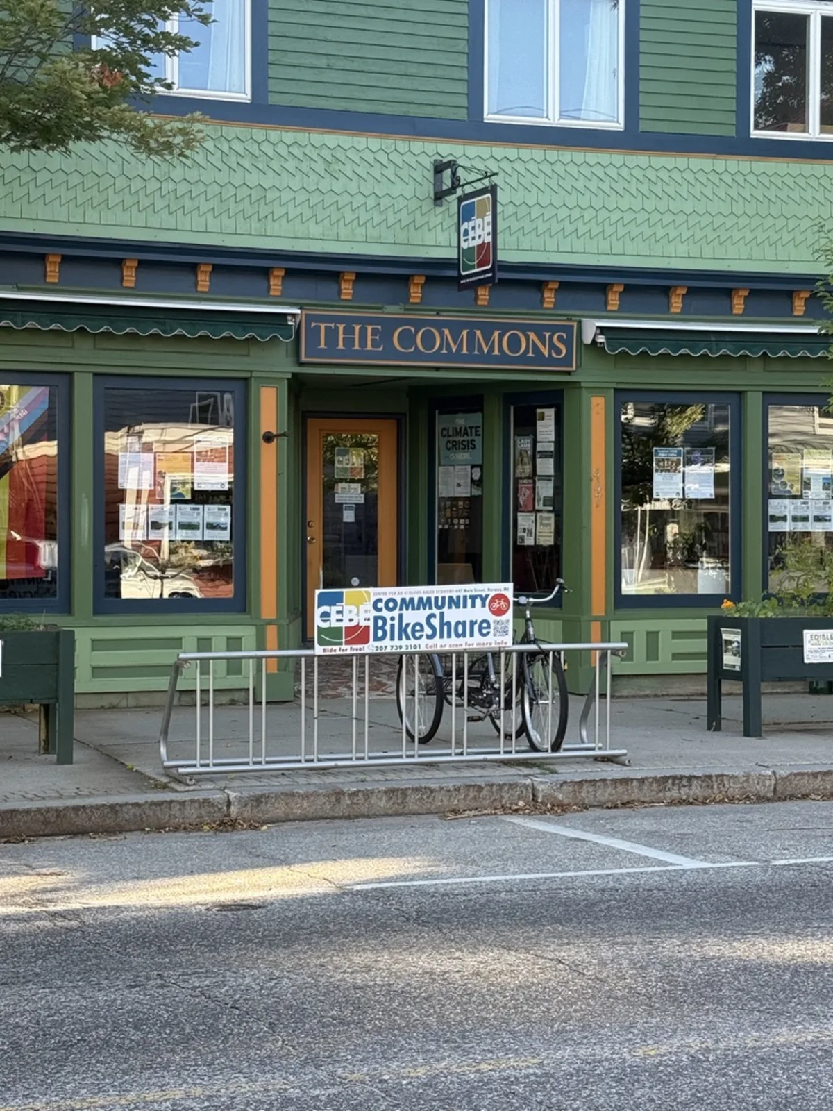 A green storefront in Norway, Maine marked THE COMMONS, with a public bike rack and a sign for the CEBE Community BikeShare reading ride for free.