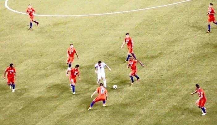 Aerial view of a football match, with an Argentine player in white surrounded by six Chilean players in orange, seen from above the pitch.