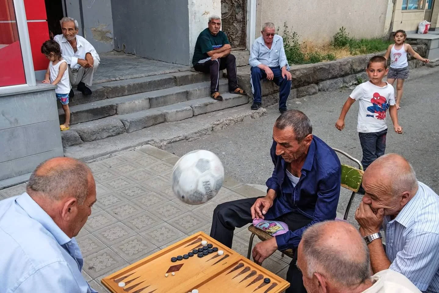 Older men playing backgammon on a chair-table in a stone alley while a soccer ball is kept aloft above them, with children watching from doorways.