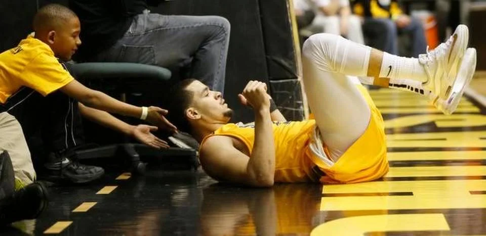 Fred VanVleet lying on the sideline floor after a hard play at Wichita State, a young ball kid in a matching jersey reaching out to greet him.
