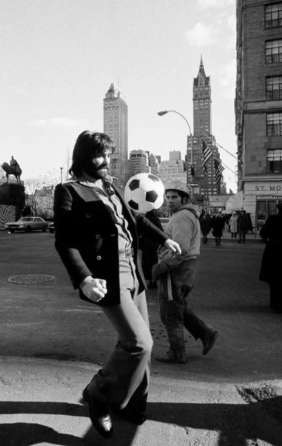 George Best juggling a soccer ball on a street corner in Manhattan, near Central Park, wearing a peacoat and jeans, with the New York skyline behind him.