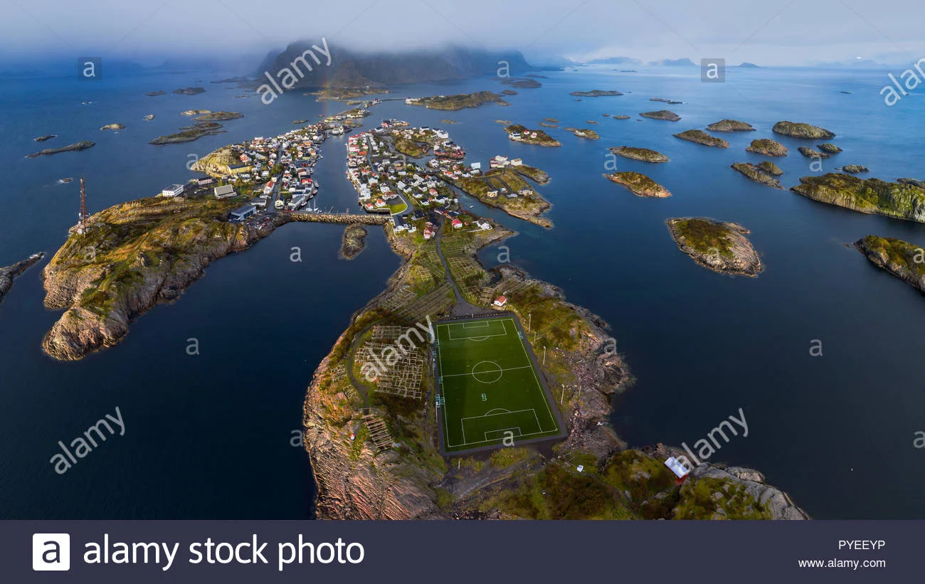 Aerial view of a full-size soccer pitch on a small rocky island in the Lofoten archipelago in northern Norway, surrounded by fjord water and misty mountains.