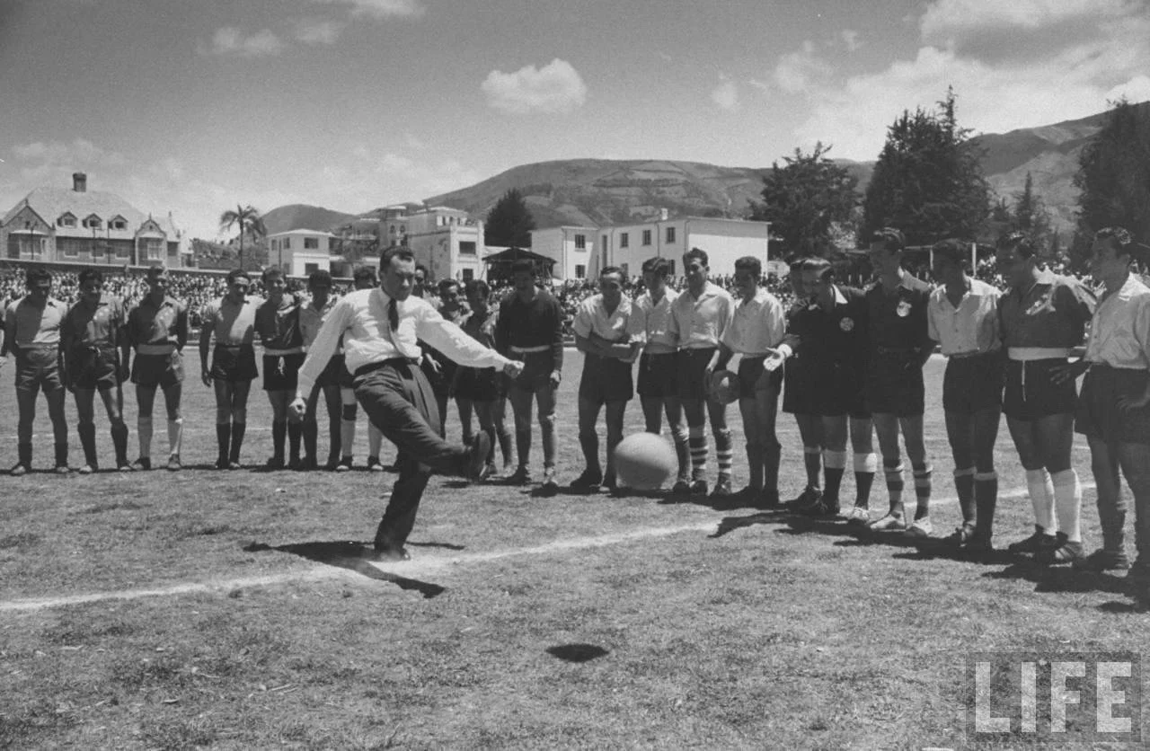 Vintage black and white LIFE Magazine photograph of a man in a suit ceremonially kicking a soccer ball with rows of players and officials watching in a stadium under mountain scenery.