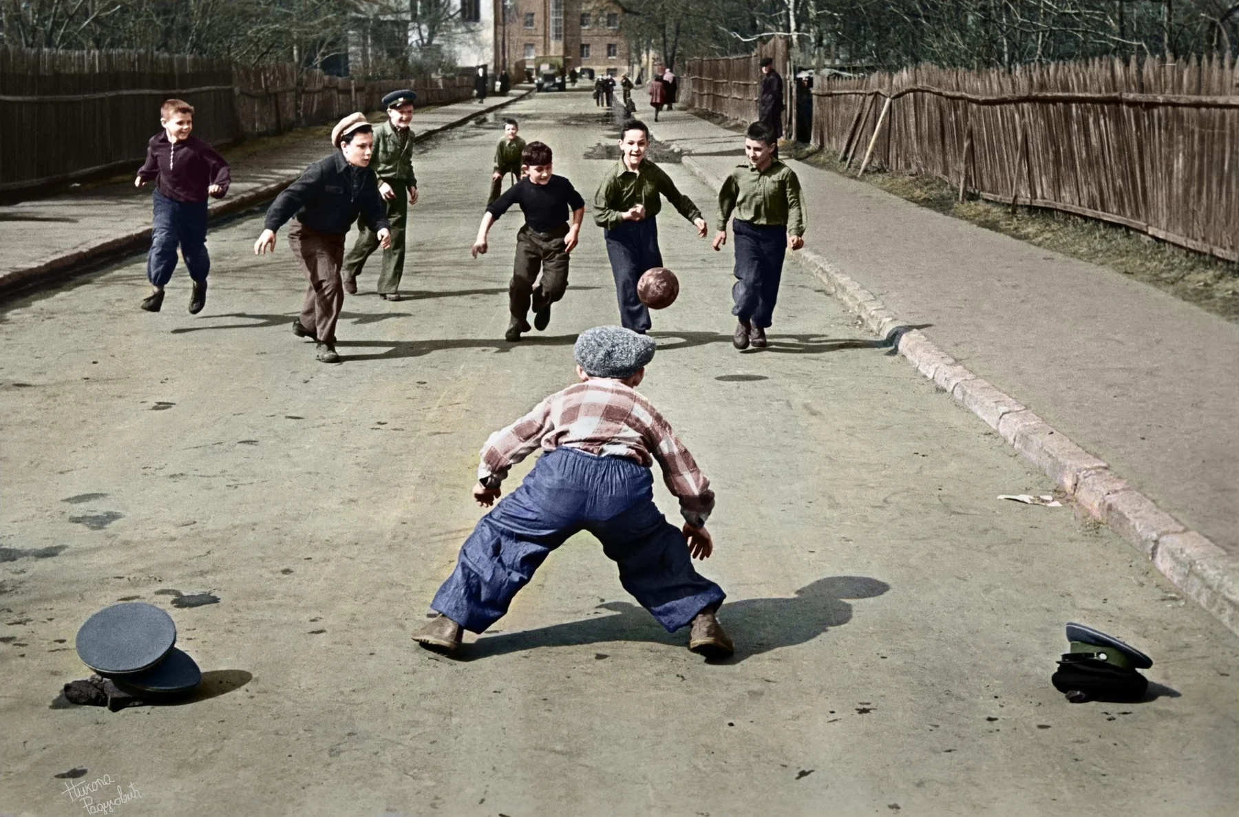 Colorized vintage photo of children playing street football on a dirt road lined with wooden fences, one boy crouched as goalkeeper between two hats used for goalposts.