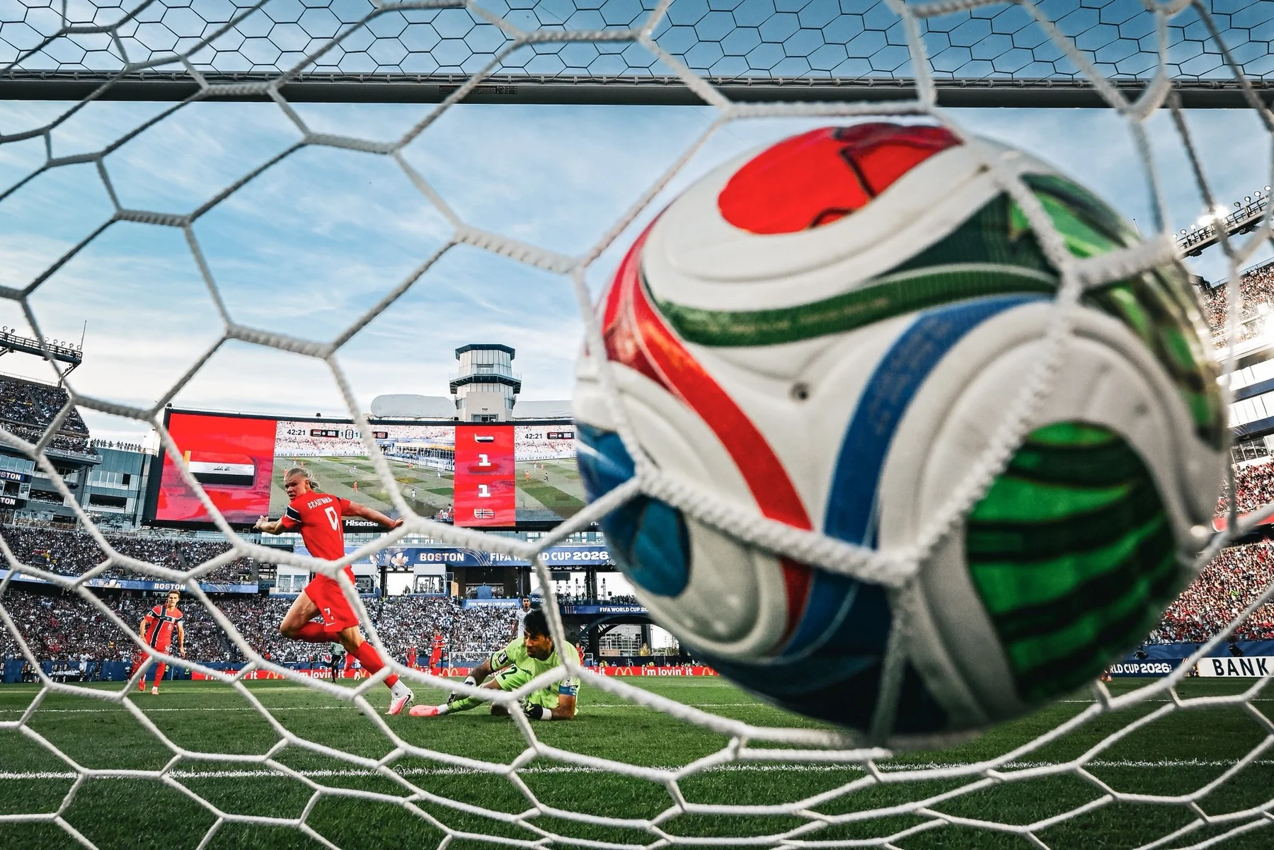 Photograph looking through the back of a soccer goal net at a World Cup 2026 match in Boston, with the ball visible in the foreground and stadium beyond.